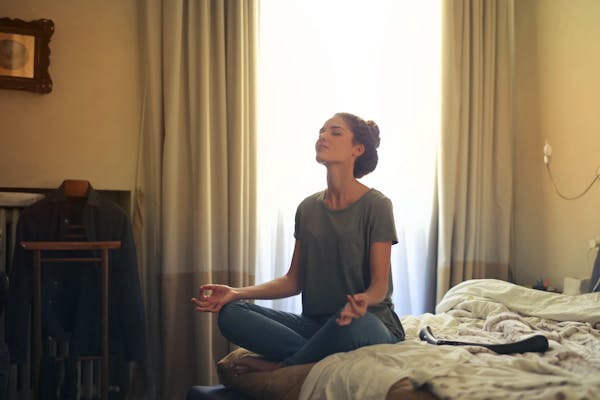 Woman meditating in a seated position on a bed, practicing mindfulness with eyes closed in a calm bedroom setting